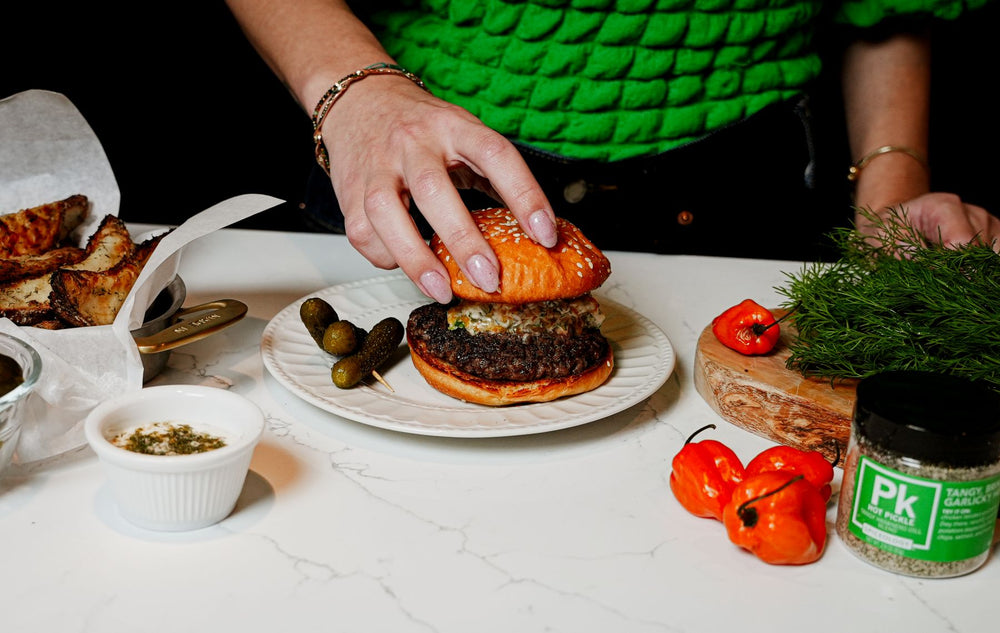 Person placing a bun on a burger beside a Spiceology PK Tangy Garlic jar, habanero peppers, and fresh dill on a marble counter