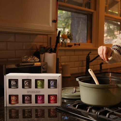 A person sprinkles spices into a green pot on a stove. In the foreground, a box labeled Spiceology holds jars of spices, each with labels resembling periodic table elements. The kitchen is warm and well-lit.