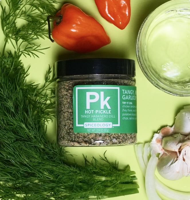 Jar of Spiceology Hot Pickle Tangy Habanero Dill Blend surrounded by dill, red peppers, and garlic on a green background
