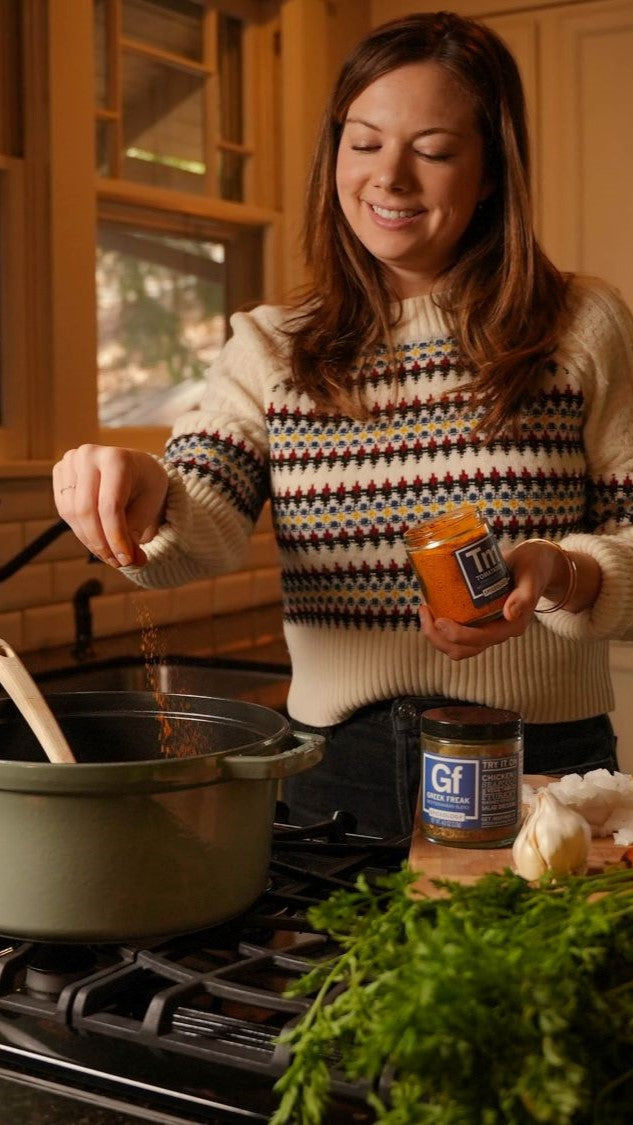 A woman cooking in a warmly lit home kitchen with Spiceology Greek Freak and Tamatador next to fresh vegetables on the counter