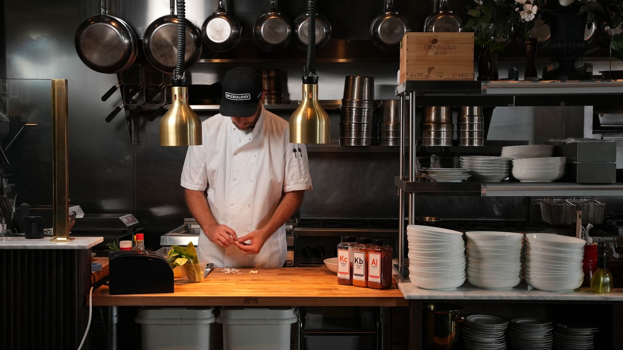 A chef in a white jacket prepping food at a wooden table in a moody restaurant kitchen with pendant lights