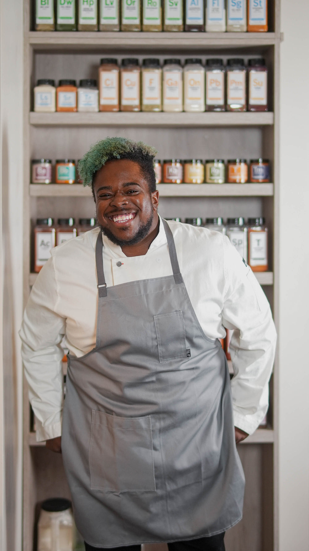 A smiling chef in a white shirt and gray apron stands in front of shelves filled with colorful spice jars.