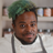 A person with short curly green hair, a trimmed beard, and wearing a white chef's jacket and gray apron smiles softly while standing indoors in front of shelves with jars.
