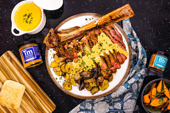 A large steak with a bone is sliced and served with roasted potatoes and mushrooms, topped with yellow sauce, on a round plate. Surrounding items include jars of seasonings, bread, a bowl of sauce, and roasted carrots.