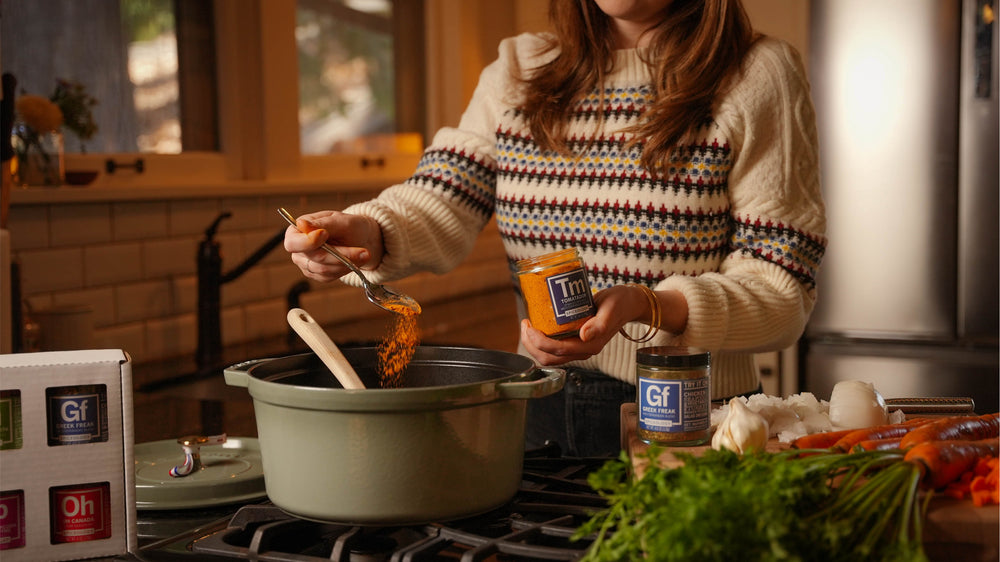 A woman in a patterned sweater adds seasoning from a jar into a green pot on a stove. Fresh herbs, garlic, and other ingredients are on the counter nearby. The kitchen is warmly lit.