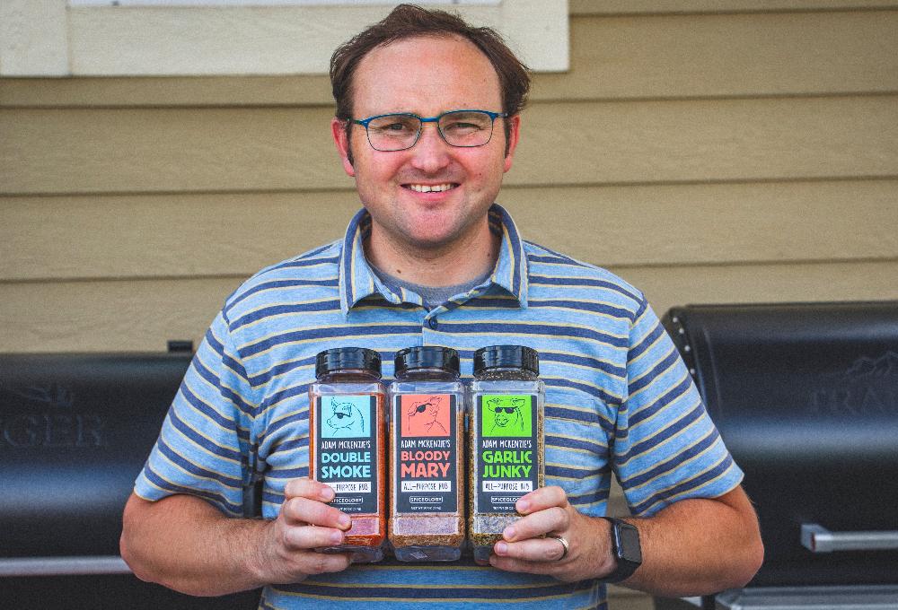 A man with glasses in a striped shirt smiling while holding multiple Spiceology blend jars