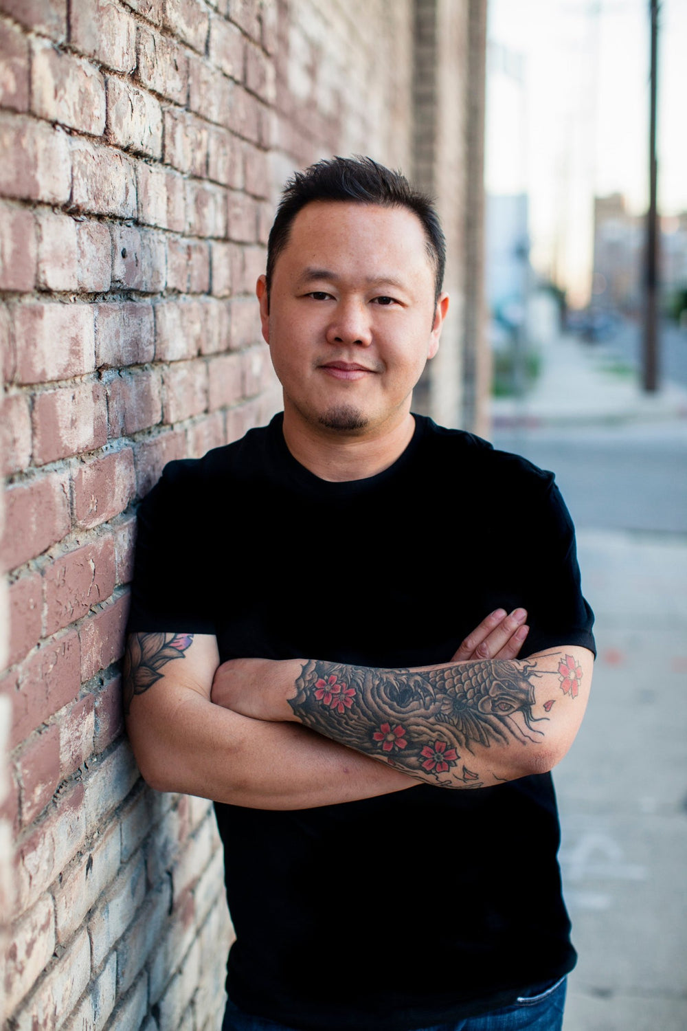 Headshot of a man with tattooed arms in a black t-shirt leaning against a brick wall