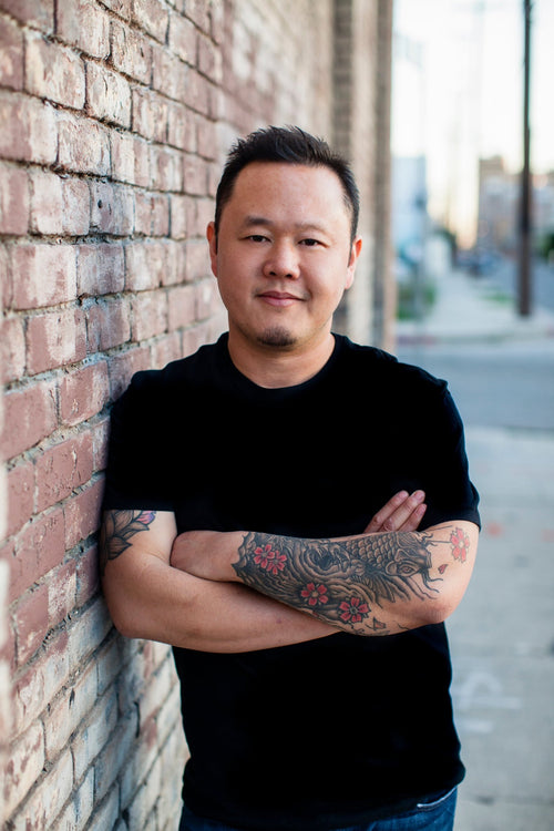 Headshot of a man with tattooed arms in a black t-shirt leaning against a brick wall