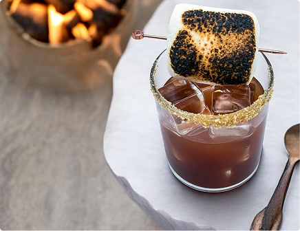 Cocktail with a flaming toasted marshmallow garnish on top in a dark glass on a white surface
