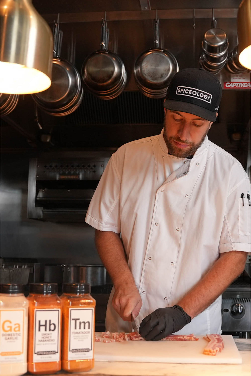 A chef in a white jacket preparing food in a professional kitchen, with an orange Spiceology blend jar visible in the foreground